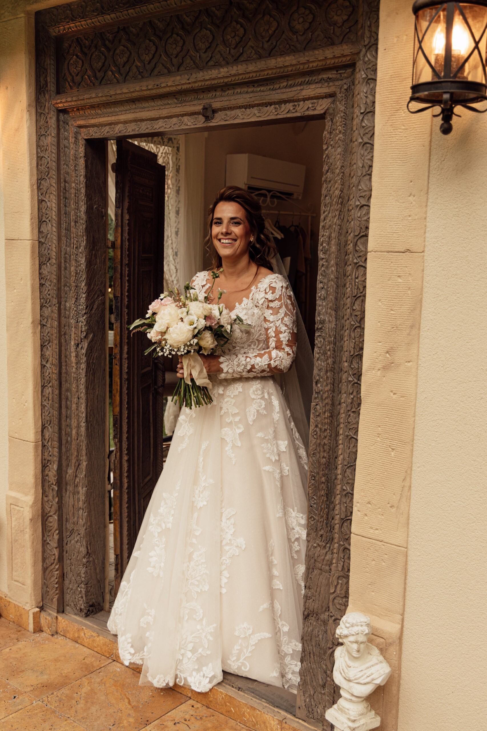 Real bride Janine in een romantische A-lijn jurk met kanten mouwen, staand in een sierlijke deuropening met haar boeket